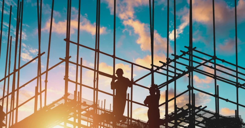 a construction site featuring scaffolding and workers in silhouette against a sunset sky